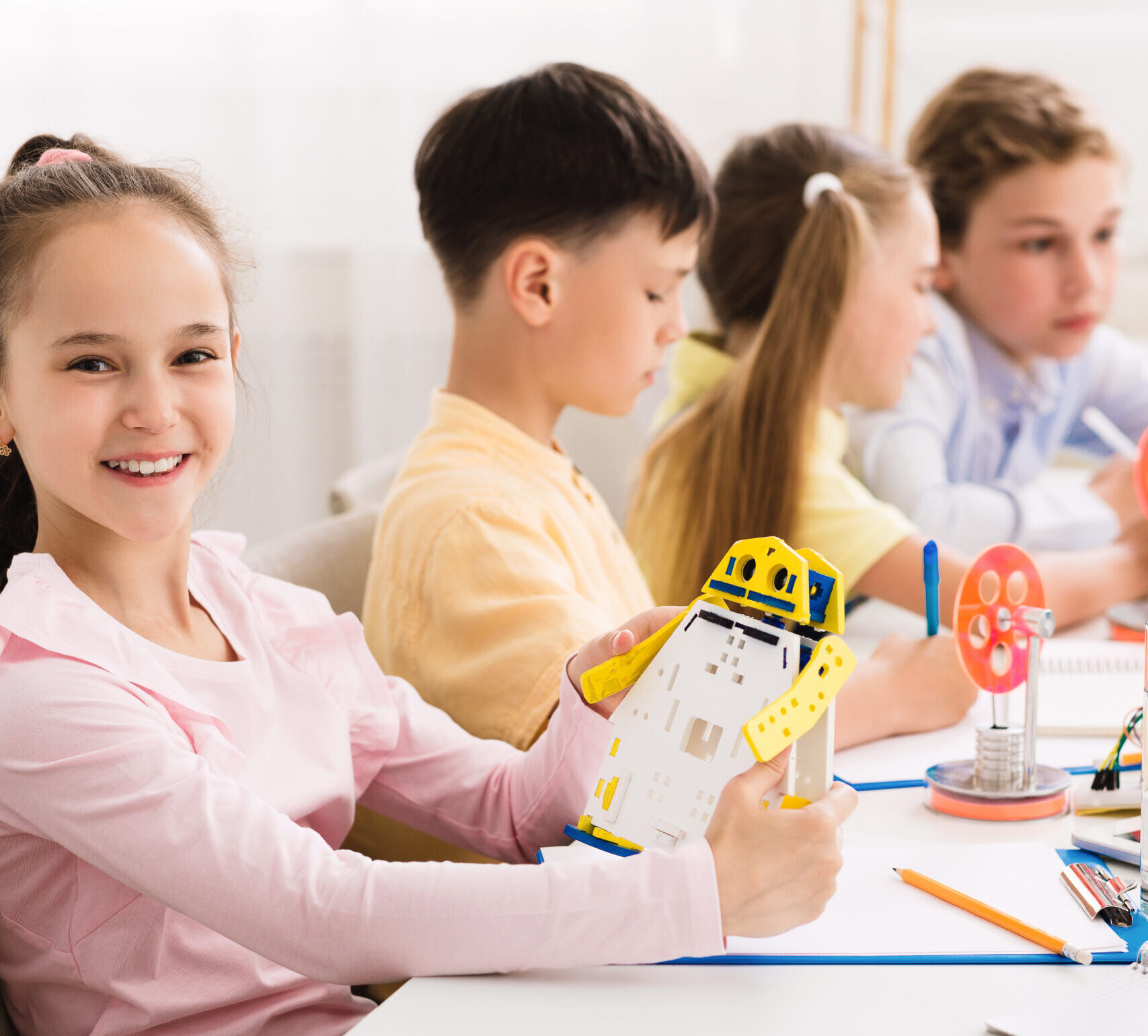 Stem education. Happy girl creating robot at classroom, smiling to camera with classmates on background